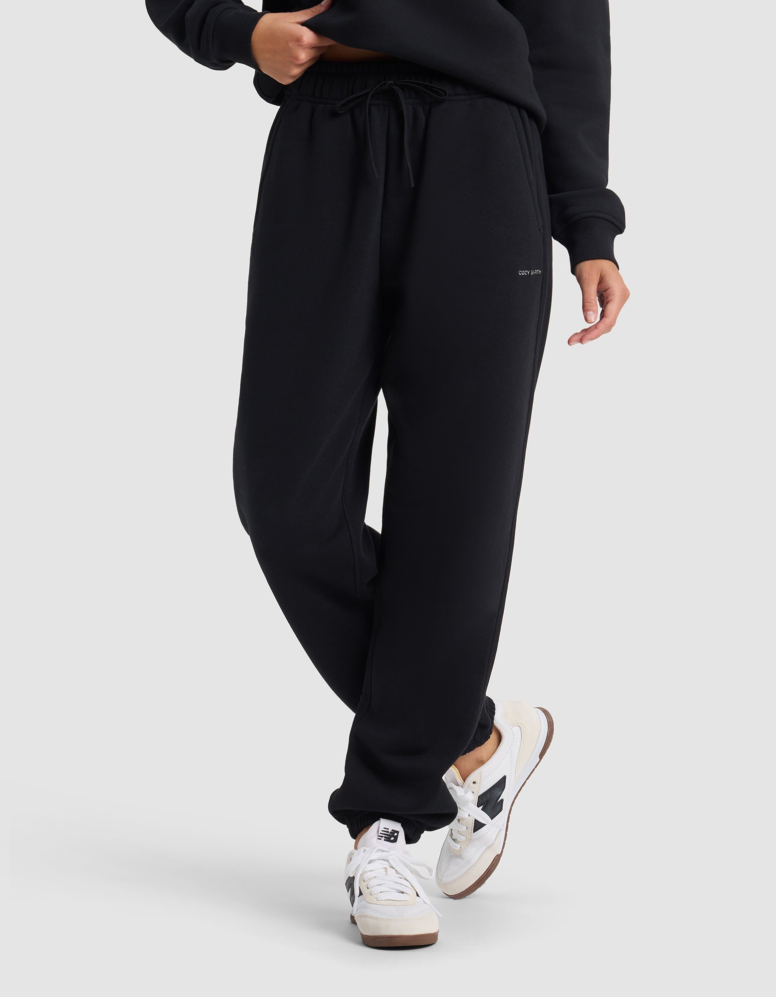 A woman wearing Cozy Earth’s Women's Cityscape 2.0 Sweatpant and a matching black sweatshirt with white sneakers featuring black accents stands against a plain light gray background.|Color:Jet Black