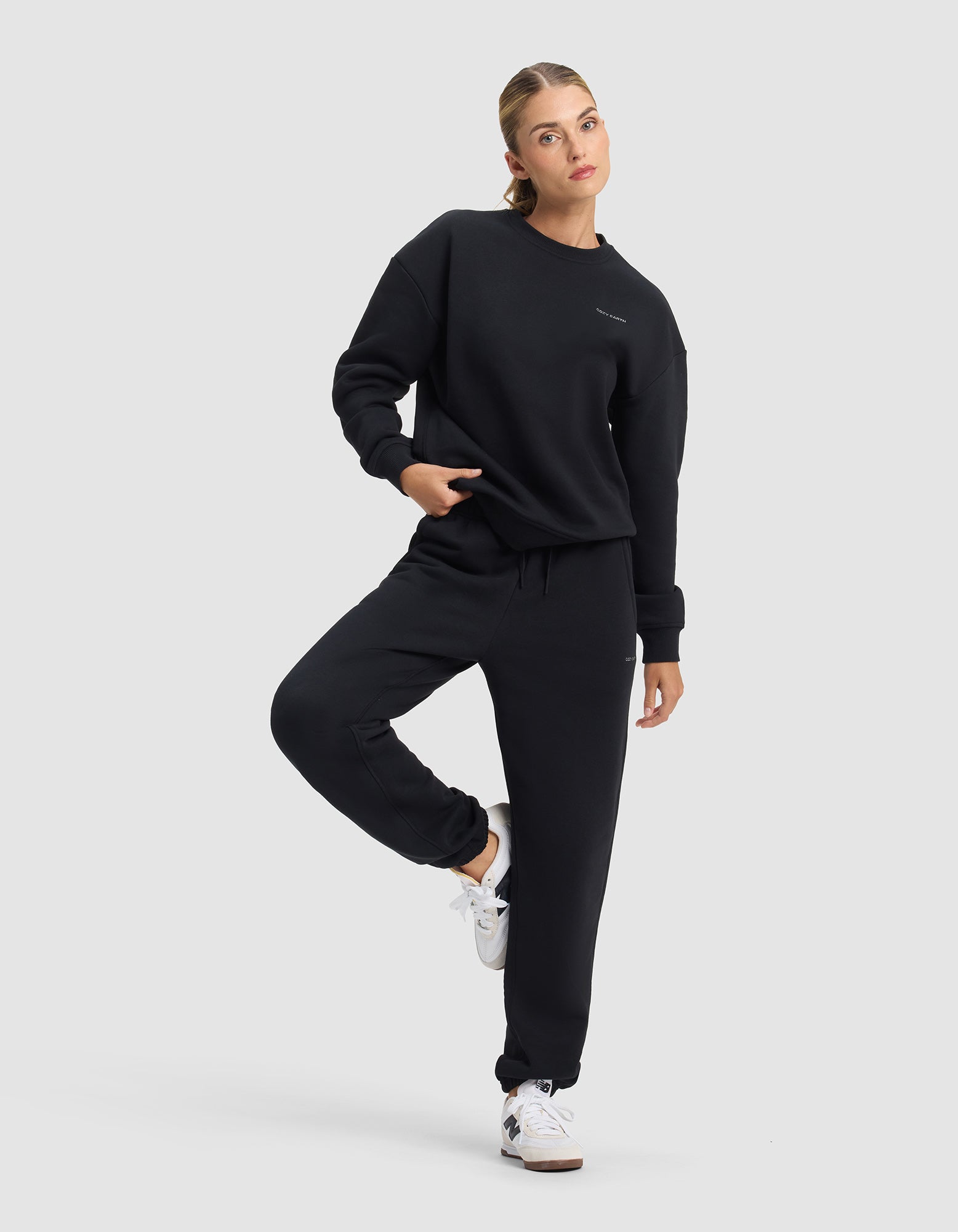 A woman stands on one leg, wearing Cozy Earth's Women's Cityscape 2.0 Sweatpant in black with a matching sweatshirt and white sneakers, looking to the side against a plain light gray background.