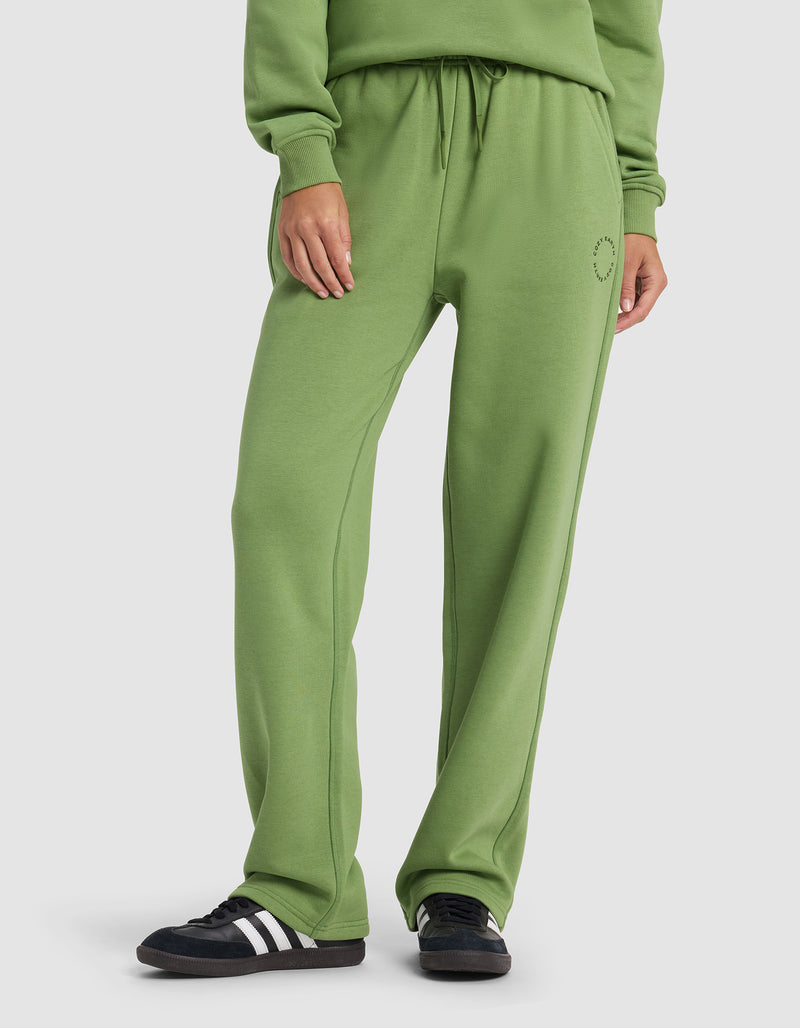 A woman wears Cozy Earth’s Women’s Cityscape 2.0 Wide Leg Pant in green with a matching sweatshirt and black sneakers with white stripes, standing against a plain white background.