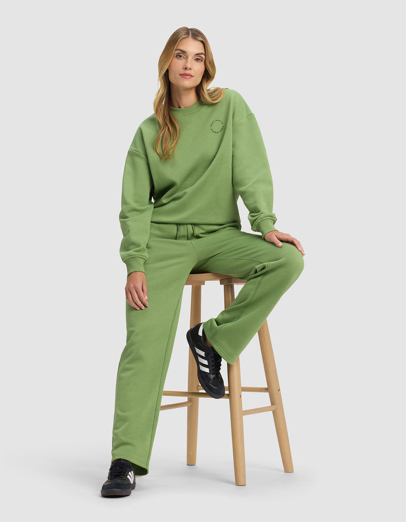 A woman with long blond hair sits on a wooden stool, wearing Cozy Earth's Women's Cityscape 2.0 Wide Leg Pant in green with a matching loose sweatshirt and black Adidas sneakers against a plain light gray background.