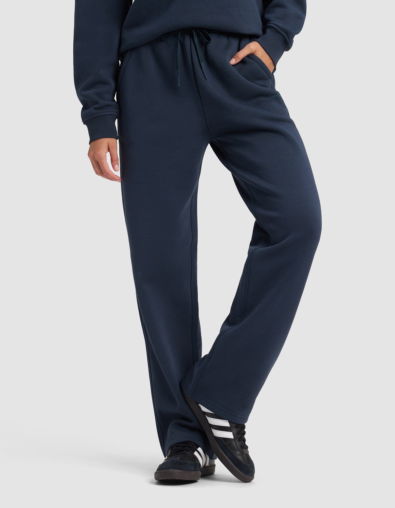 A woman wears Cozy Earth's Women's Cityscape 2.0 Wide Leg Pant in navy with a matching sweatshirt and black sneakers with white stripes, standing on a white background with one leg crossed in front of the other.|Color:Eclipse