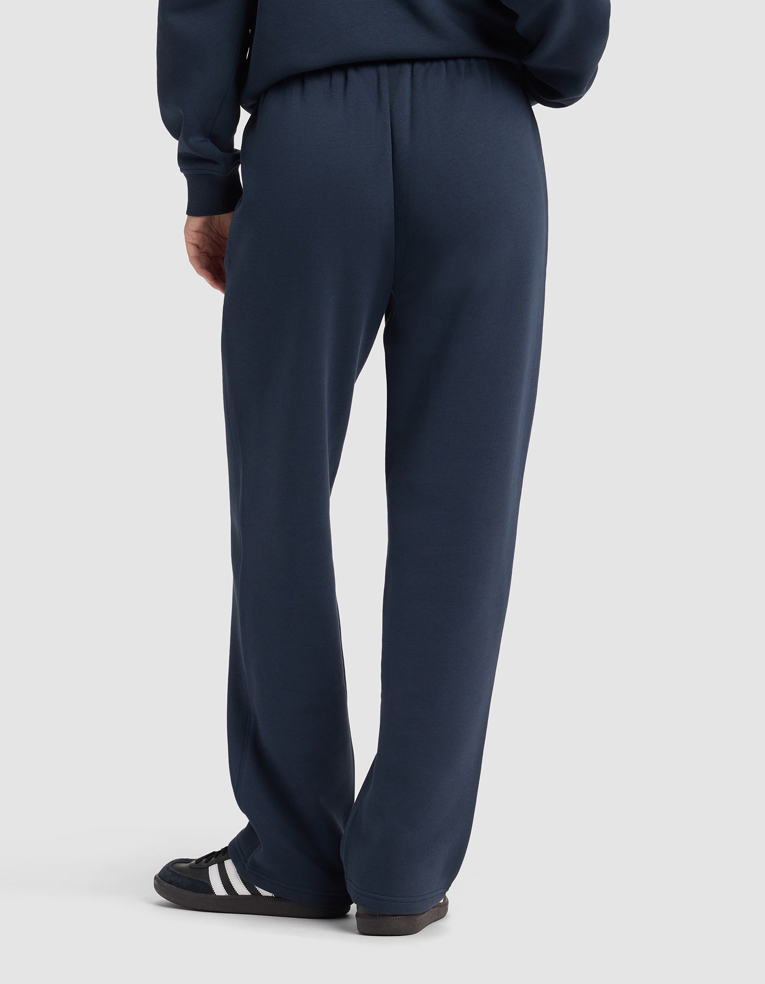 A woman wearing Cozy Earth’s Women's Cityscape 2.0 Wide Leg Pant in navy, paired with black sneakers with white stripes, stands with her back to the camera against a plain light grey background.