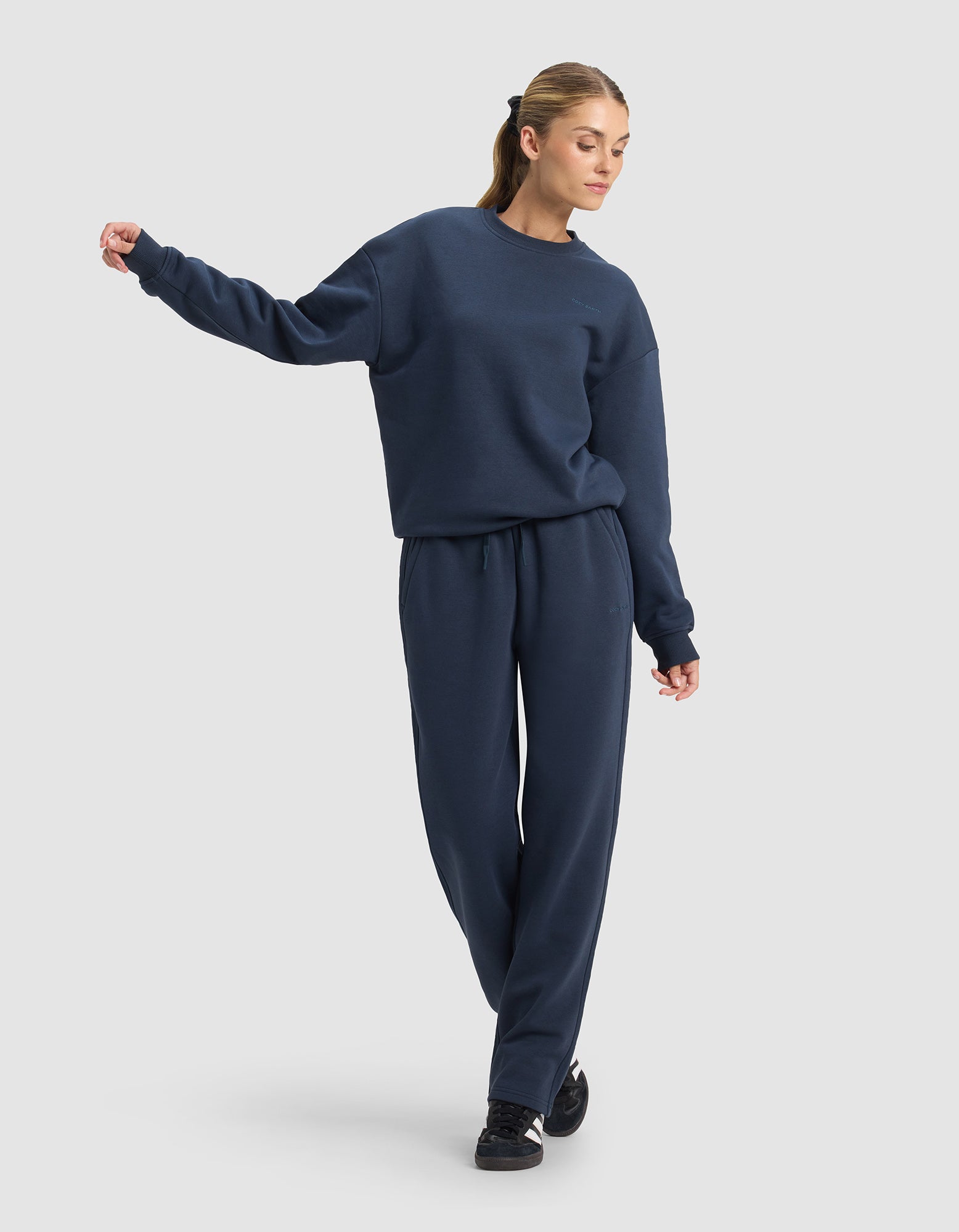A woman in Cozy Earth’s Women’s Cityscape 2.0 Wide Leg Pant and a matching navy sweatshirt stands with one arm raised, looking to the side. She wears black sneakers, her light brown hair tied back, against a plain light gray background.