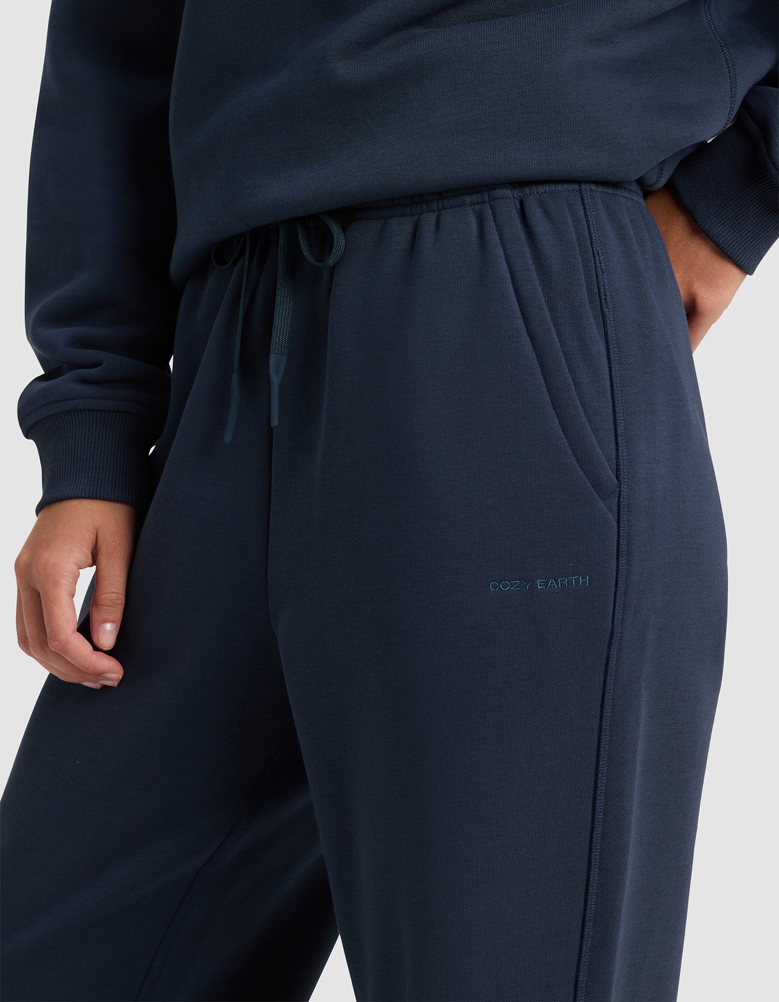 A person wears navy blue Cozy Earth Women's Cityscape 2.0 Wide Leg Pants and a matching sweatshirt, with one hand in a pocket. The pants feature an elastic drawstring waistband and "COZY EARTH" embroidered near the pocket.