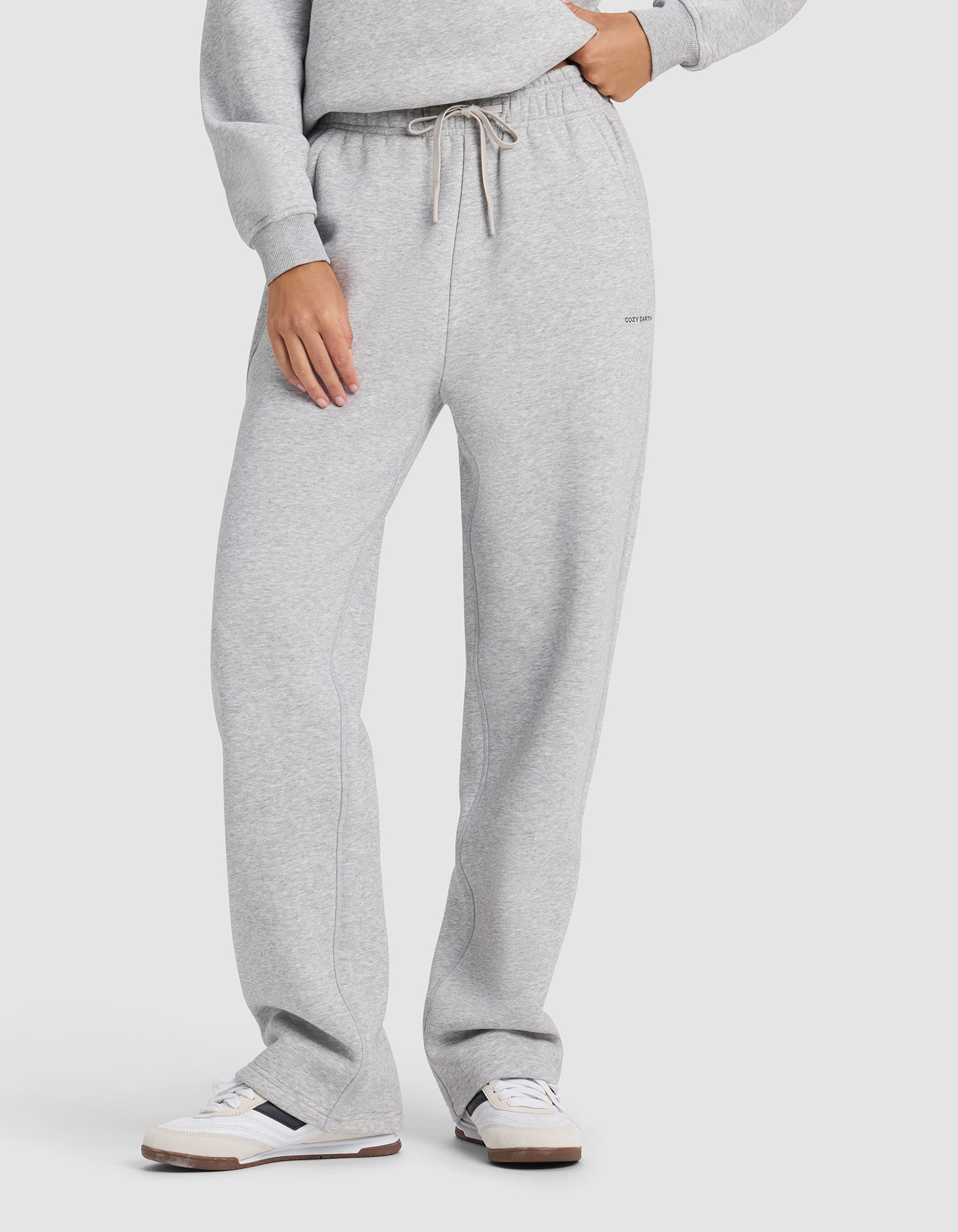 A woman models Cozy Earth's Women's Cityscape 2.0 Wide Leg Pant in light gray, paired with a matching sweatshirt and white sneakers with light brown soles. She stands against a plain white background, one hand in her pocket.