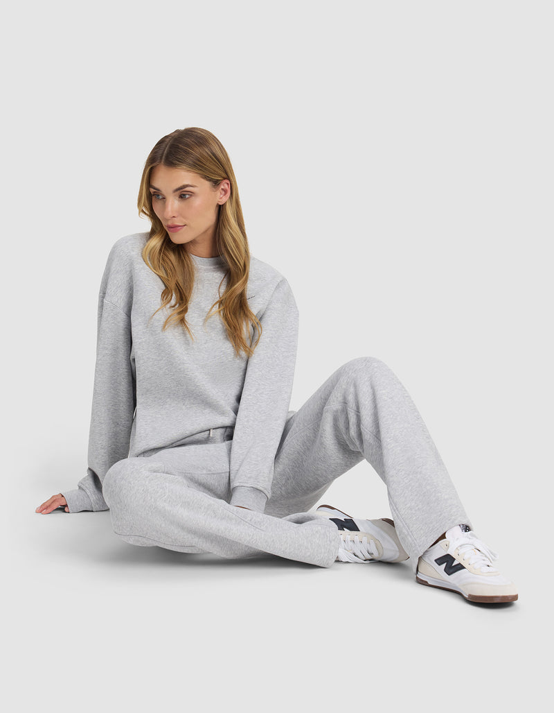 A woman with long, light brown hair sits on the floor wearing Cozy Earth's Women's Cityscape 2.0 Wide Leg Pant in light gray, paired with a matching sweatshirt and white sneakers with black details against a plain background.