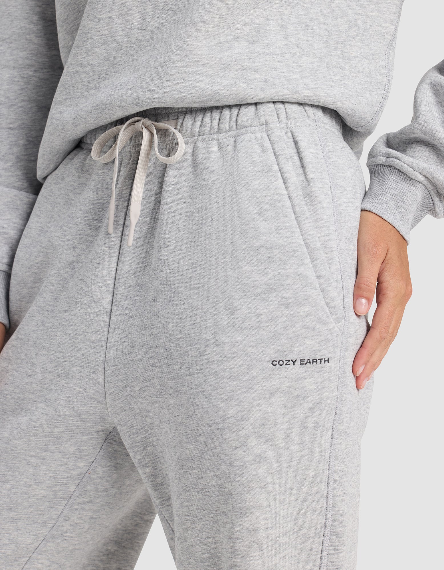 A person wears Cozy Earth Women's Cityscape 2.0 Wide Leg Pants in light gray with a drawstring waist and “COZY EARTH” branding near the pocket, paired with a matching sweatshirt and their hand in the sweatpants pocket.|Color:Heather Grey