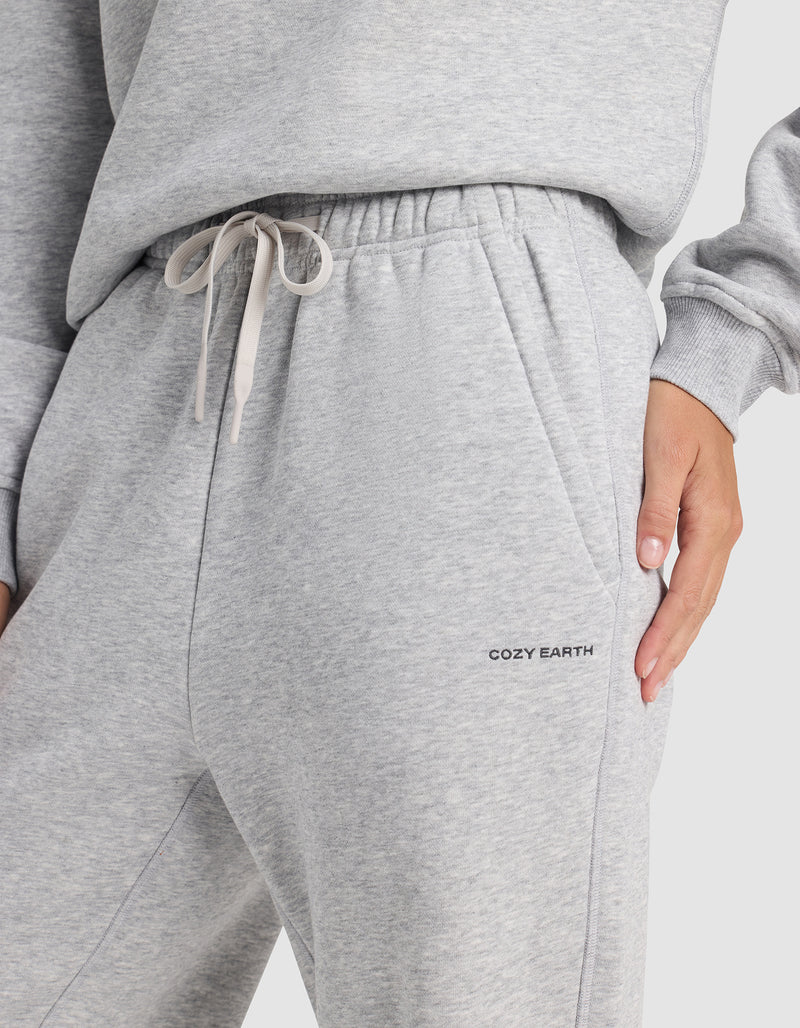 A person wears Cozy Earth Women's Cityscape 2.0 Wide Leg Pants in light gray with a drawstring waist and “COZY EARTH” branding near the pocket, paired with a matching sweatshirt and their hand in the sweatpants pocket.