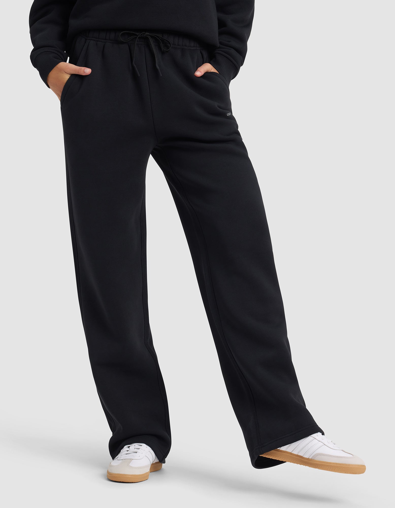 A woman wears Cozy Earth's Women's Cityscape 2.0 Wide Leg Pant in black with hands in pockets and white sneakers, standing against a light gray background. Only her lower body is visible.|Color:Jet Black