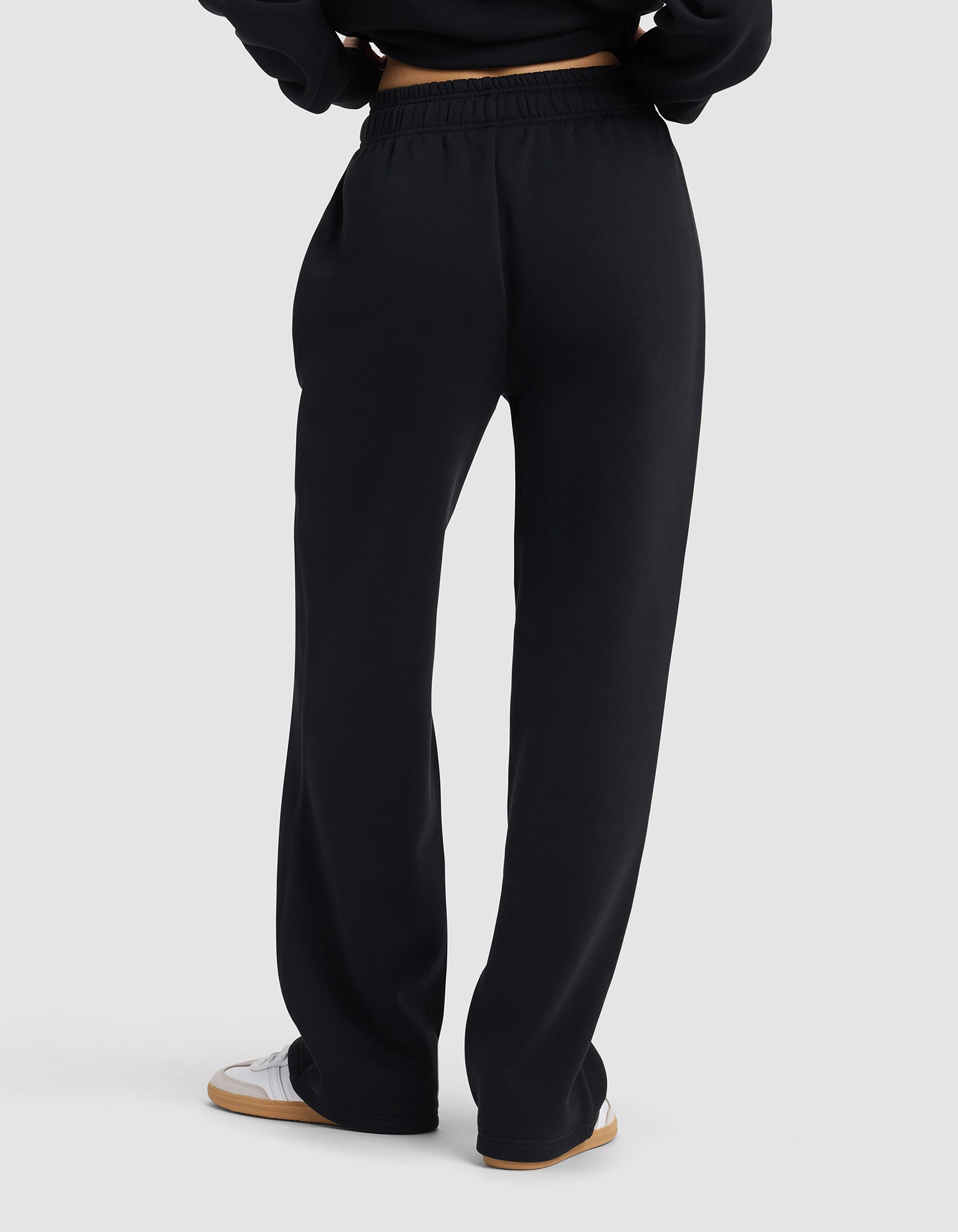 A woman wearing Cozy Earth's Women's Cityscape 2.0 Wide Leg Pant in black and white sneakers stands with her back to the camera against a plain light gray background.|Color:Jet Black