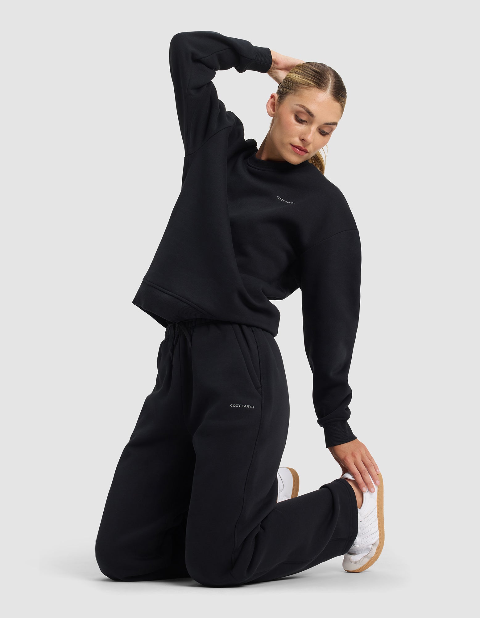 A woman with blonde hair tied back kneels on a plain gray floor, stretching in Cozy Earth's Women's Cityscape 2.0 Wide Leg Pant and a black sweatshirt, paired with white sneakers.|Color:Jet Black