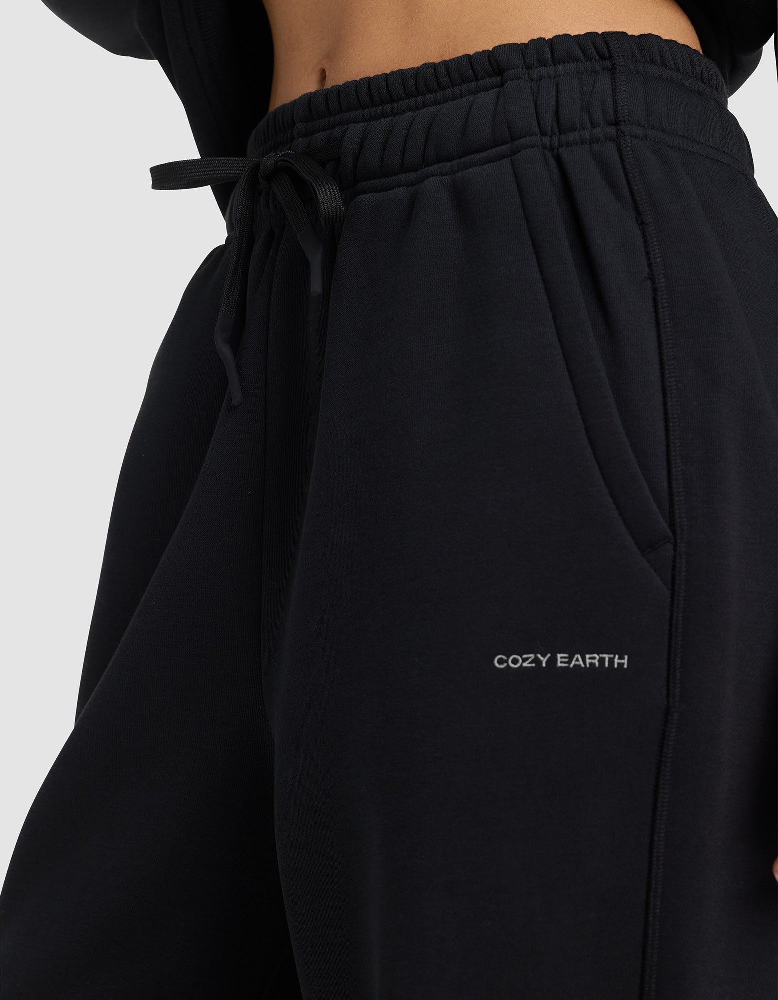 Close-up of a person wearing Cozy Earth's Women's Cityscape 2.0 Wide Leg Pant in black, featuring an elastic waistband, drawstring, and "COZY EARTH" embroidered in white on the left pocket. The midriff and hand in pocket are visible.|Color:Jet Black