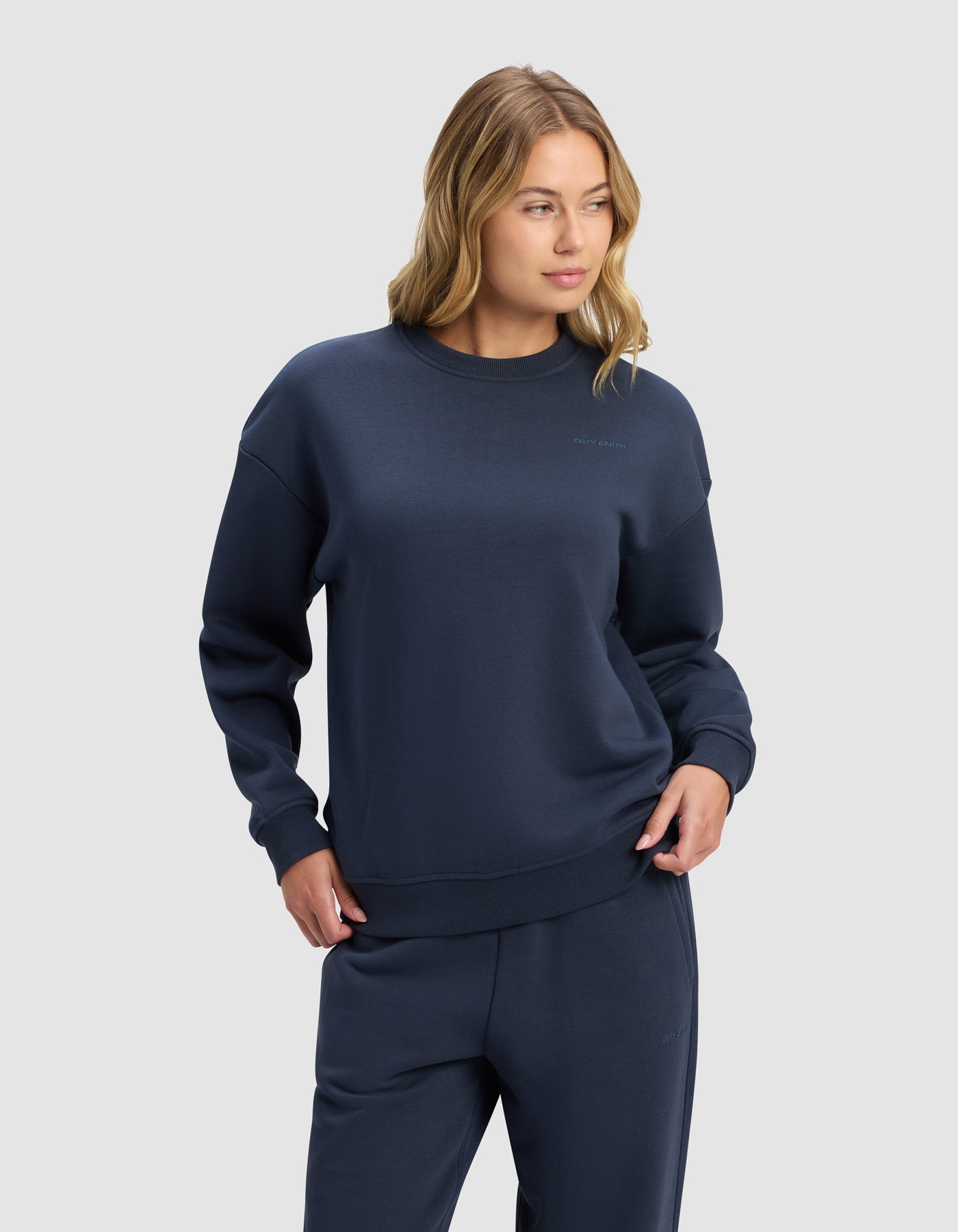 A woman with loose blonde waves wears the Cozy Earth Women's Cityscape 2.0 Crewneck in dark blue, paired with matching sweatpants, standing confidently against a light gray background.|Color:Eclipse