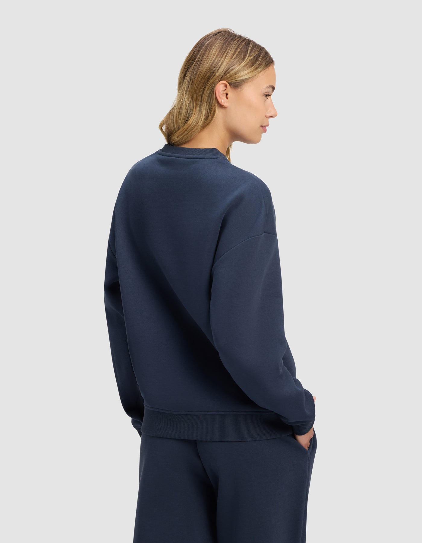 A woman with blonde hair models the Cozy Earth Women's Cityscape 2.0 Crewneck in dark blue, paired with matching pants, standing against a light gray background, hands in pockets and looking to the side.|Color:Eclipse