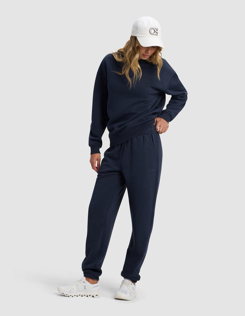 A model in the Cozy Earth Women's Cityscape 2.0 Crewneck and matching sweatpants, styled with white sneakers and a white baseball cap, stands against a light gray background, looking down with one hand in her pocket.