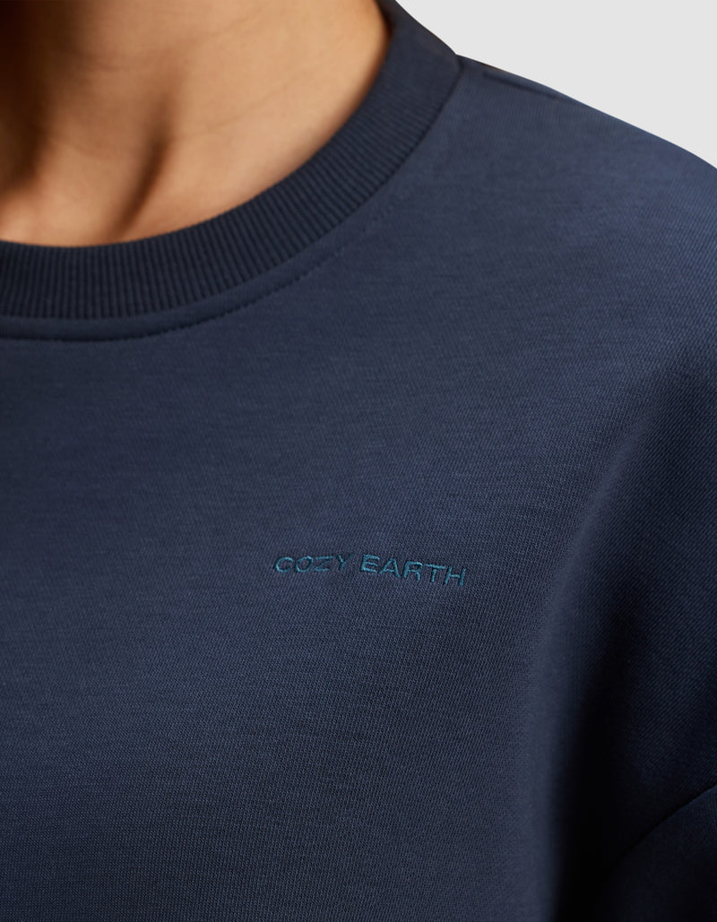 Close-up of a person wearing the Women's Cityscape 2.0 Crewneck by Cozy Earth in navy blue, featuring small light blue "Cozy Earth" embroidery on the chest. The collar and soft fabric texture are clearly visible.