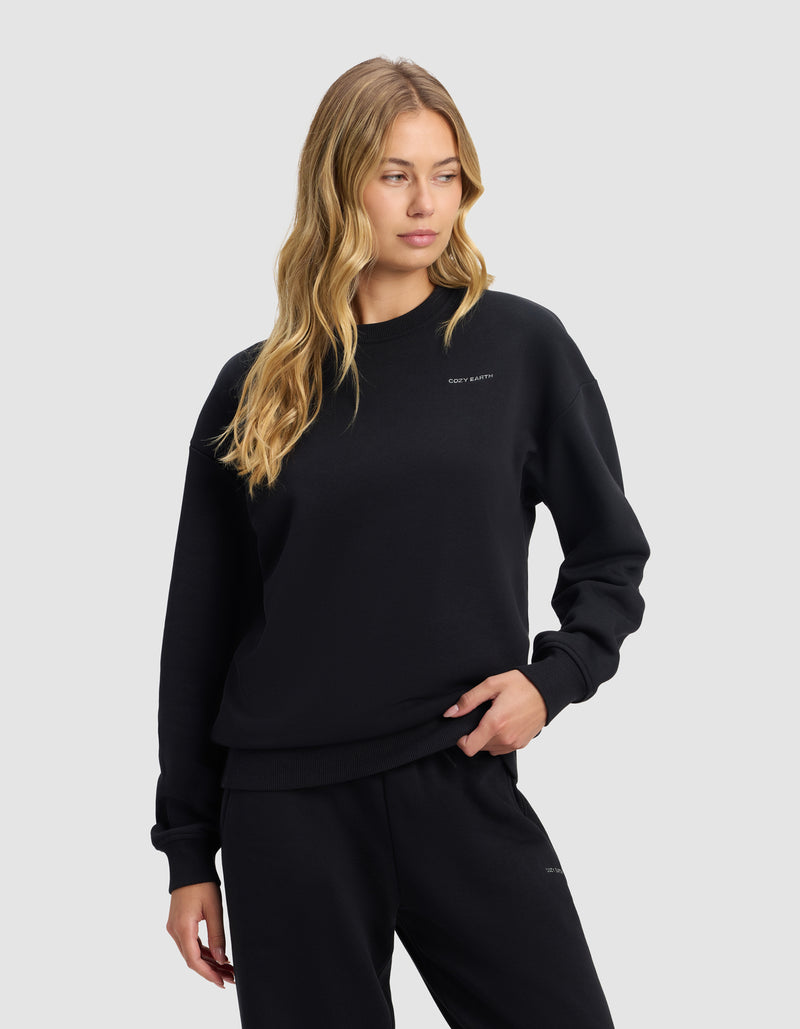 A woman with long blonde hair models the Cozy Earth Women's Cityscape 2.0 Crewneck in black, paired with matching pants, against a plain light gray background. She gazes slightly right with one hand on her hip.