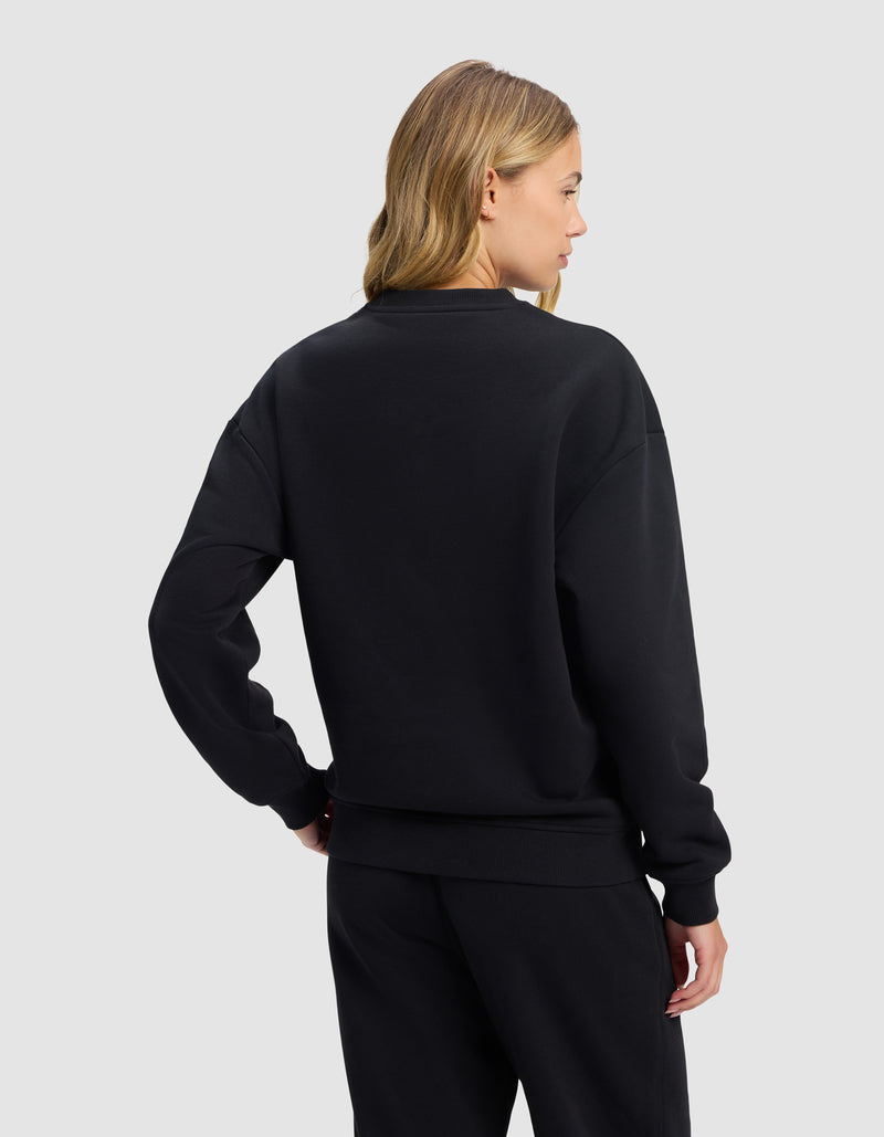 A woman with blonde hair is seen from behind wearing the Cozy Earth Women's Cityscape 2.0 Crewneck in black, paired with black pants, standing against a plain light gray background.
