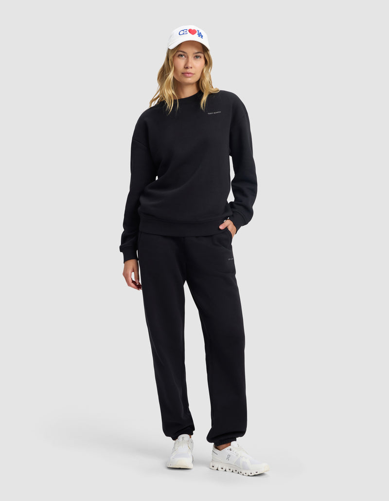 A woman models the Cozy Earth Women's Cityscape 2.0 Crewneck in black, paired with matching sweatpants, a white baseball cap, and white sneakers, standing against a gray background with one hand in her pocket.