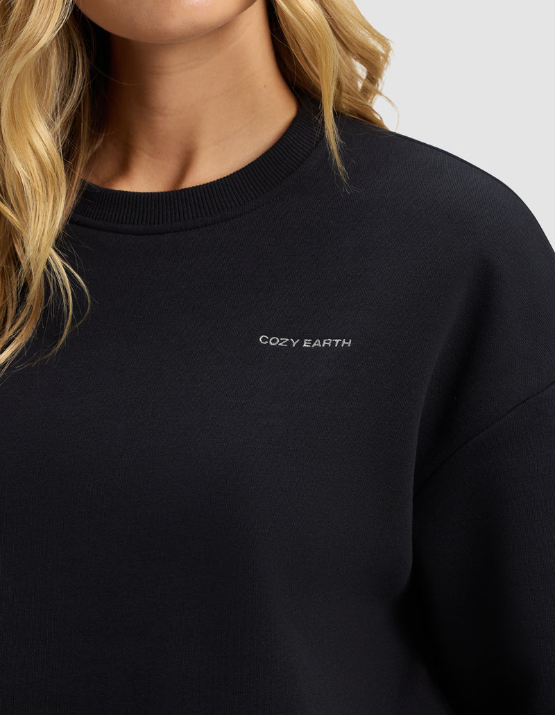 A close-up shows a person in the Women's Cityscape 2.0 Crewneck by Cozy Earth, featuring "COZY EARTH" embroidered in small white letters on the black chest; their blonde hair is visible, with most of their face out of frame.