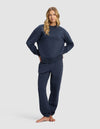A woman with long blonde hair stands barefoot in the Cozy Earth Women's Cityscape 2.0 Crewneck & Sweatpant Set, posing with hands on hips against a plain light gray background.|Color:Eclipse