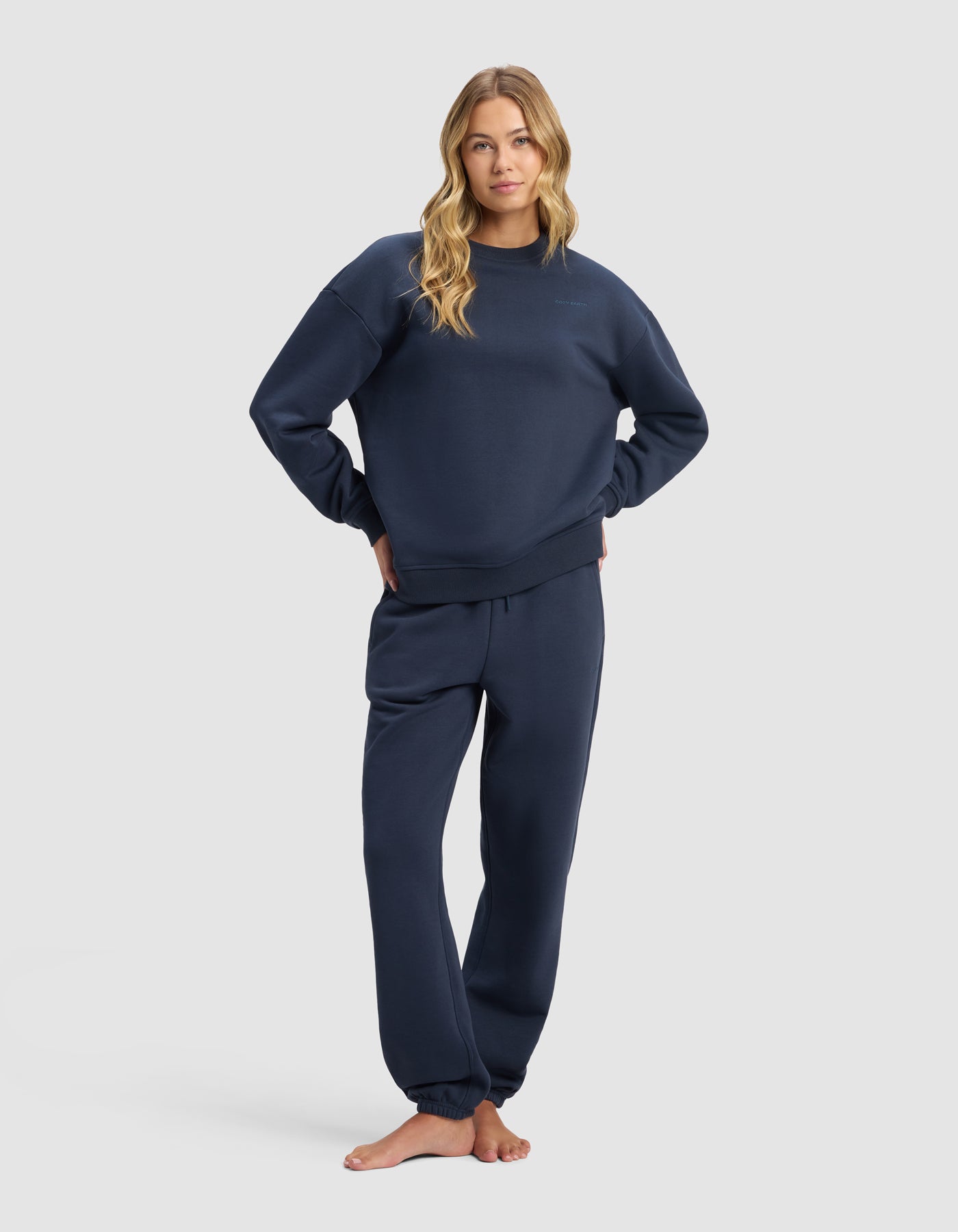 A woman with long blonde hair stands barefoot in the Cozy Earth Women's Cityscape 2.0 Crewneck & Sweatpant Set, posing with hands on hips against a plain light gray background.|Color:Eclipse