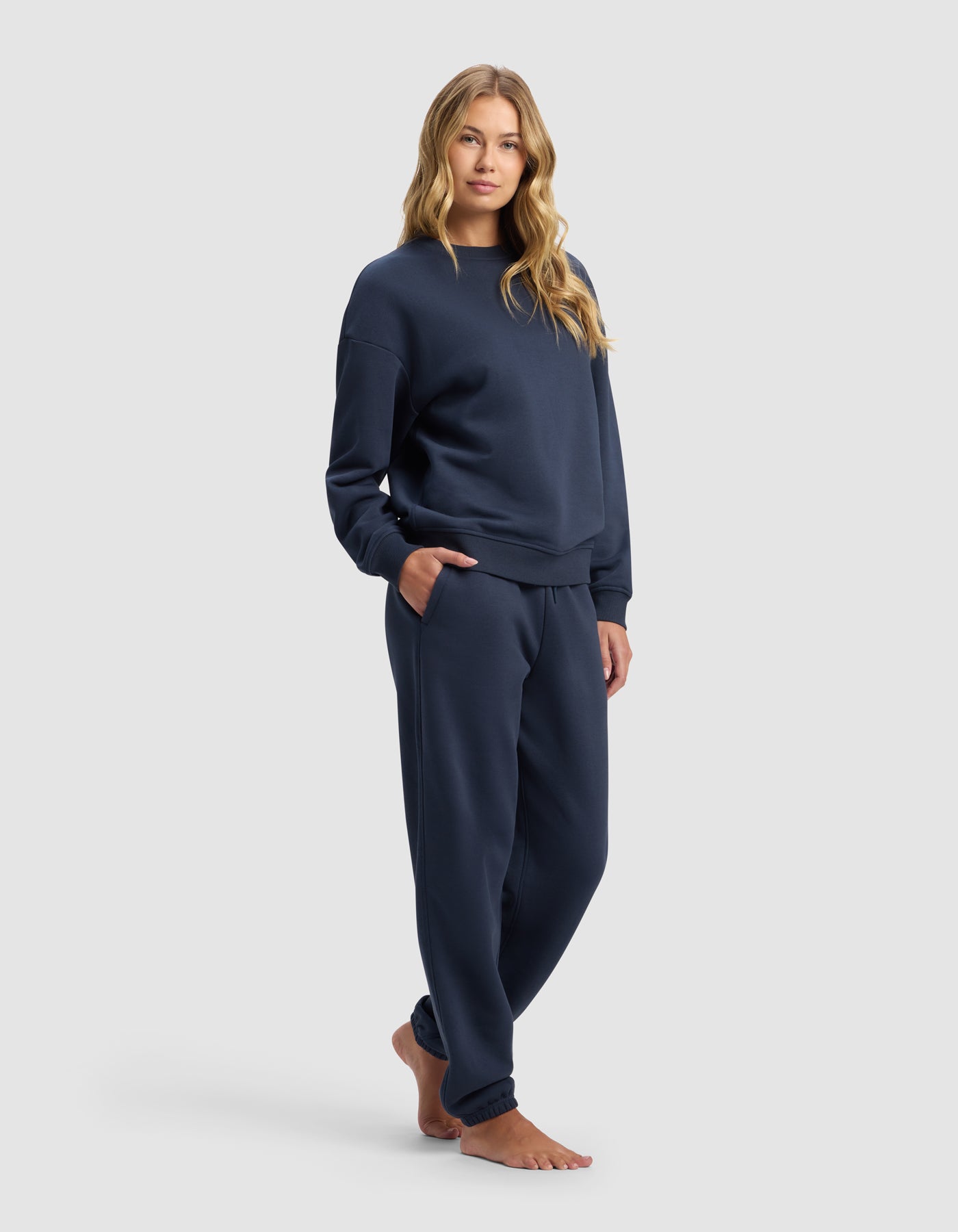 A woman with long, wavy blonde hair stands barefoot against a white background, wearing the Cozy Earth Women's Cityscape 2.0 Crewneck & Sweatpant Set in navy, hands in her pockets and a relaxed expression.|Color:Eclipse