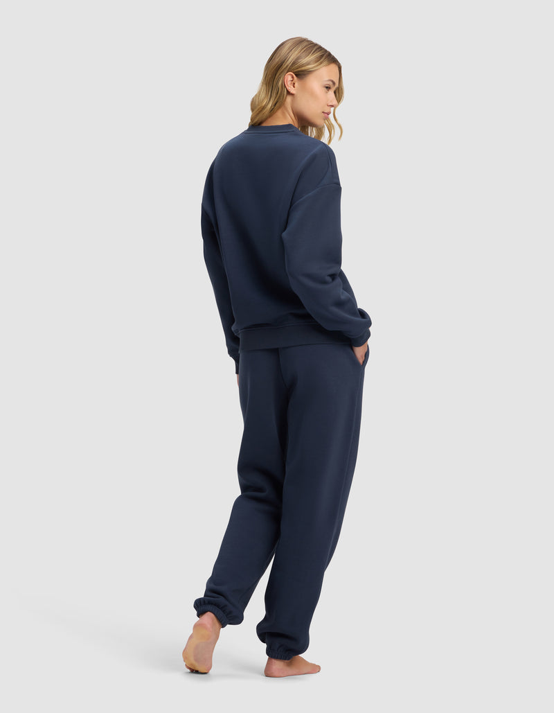 A woman stands barefoot on a light background, wearing the Cozy Earth Women's Cityscape 2.0 Crewneck & Sweatpant Set in navy blue. She faces away, glancing over her shoulder with one hand in her pocket.