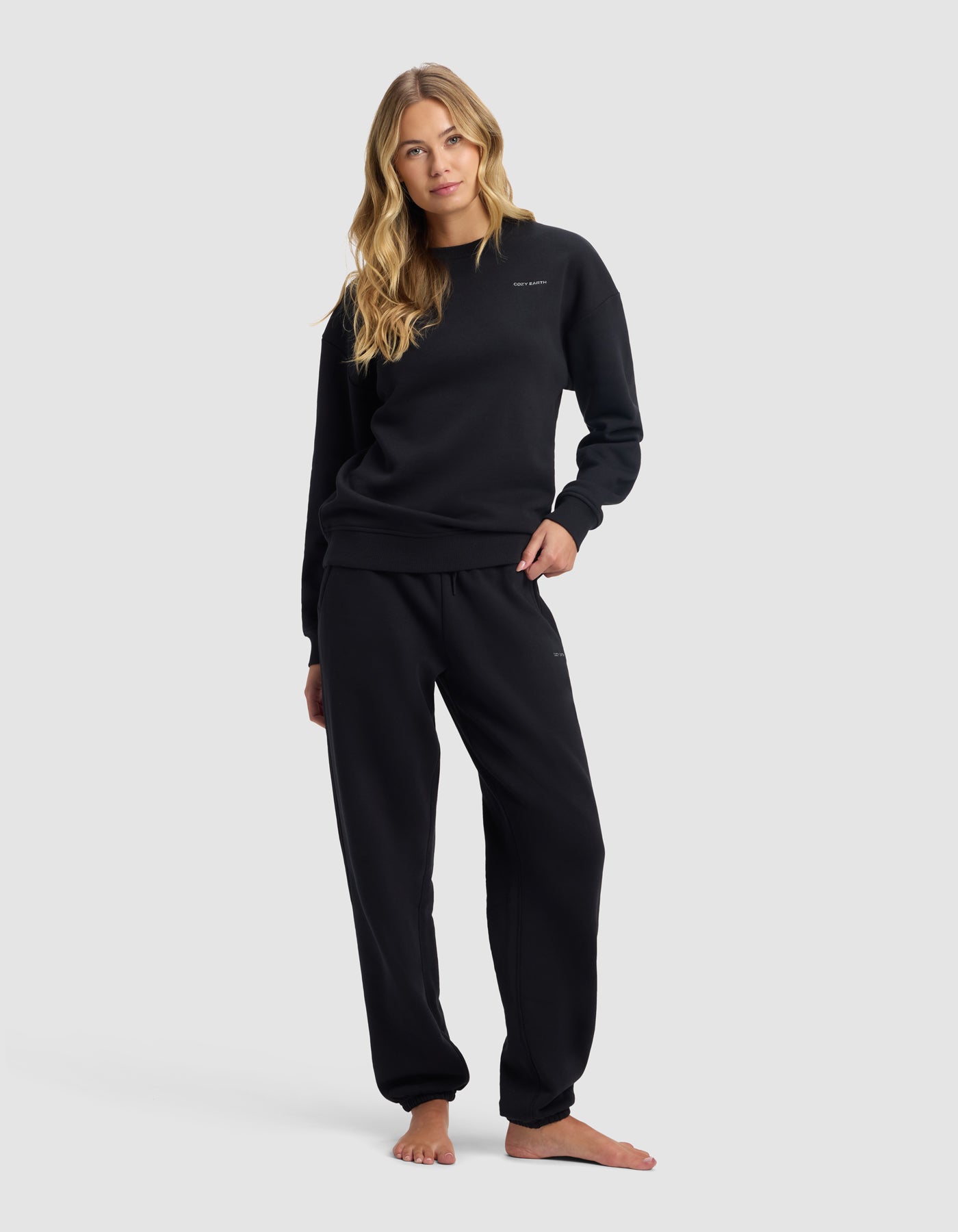 A woman with long blonde hair models the Cozy Earth Women's Cityscape 2.0 Crewneck & Sweatpant Set in black, standing barefoot with one hand in her pocket and a relaxed look against a plain light gray background.