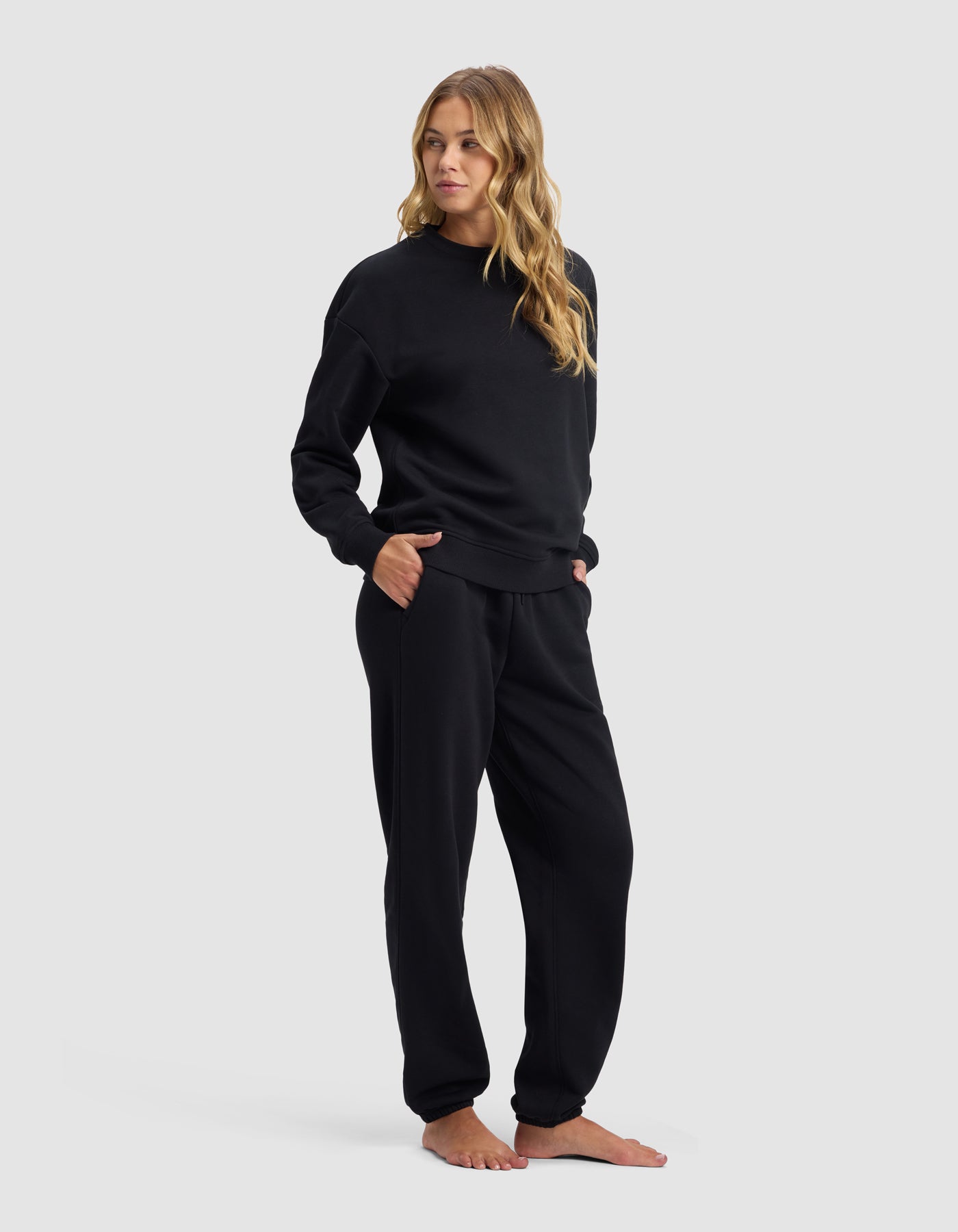 A woman with long, wavy blonde hair stands barefoot against a light gray background, wearing the Cozy Earth Women's Cityscape 2.0 Crewneck & Sweatpant Set in black with her hands in her pockets.