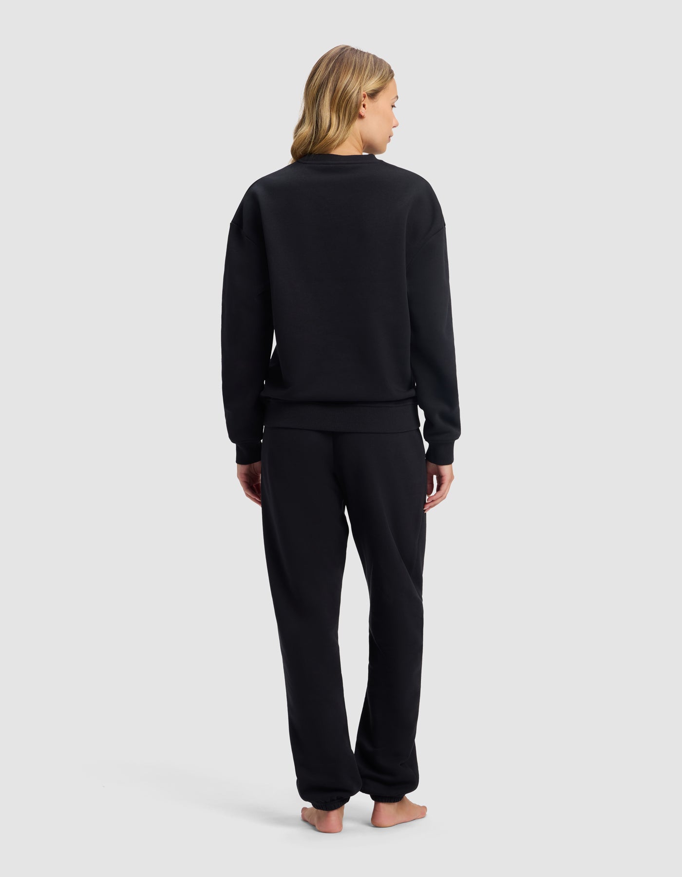 A person with long blonde hair stands barefoot, facing away, wearing the Cozy Earth Women's Cityscape 2.0 Crewneck & Sweatpant Set in black against a plain light gray background.