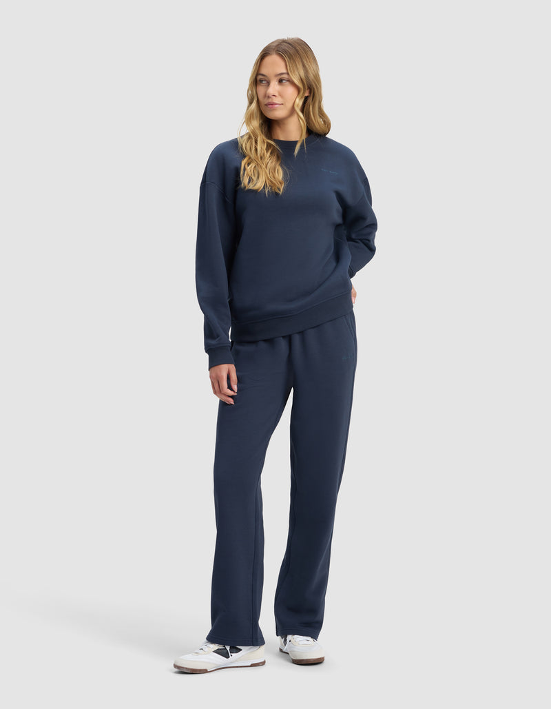 A woman with long, wavy blonde hair stands against a plain background, wearing the Cozy Earth Women's Cityscape 2.0 Crewneck & Wide Leg Pant Set in navy blue with white sneakers, looking relaxed and slightly to the side.