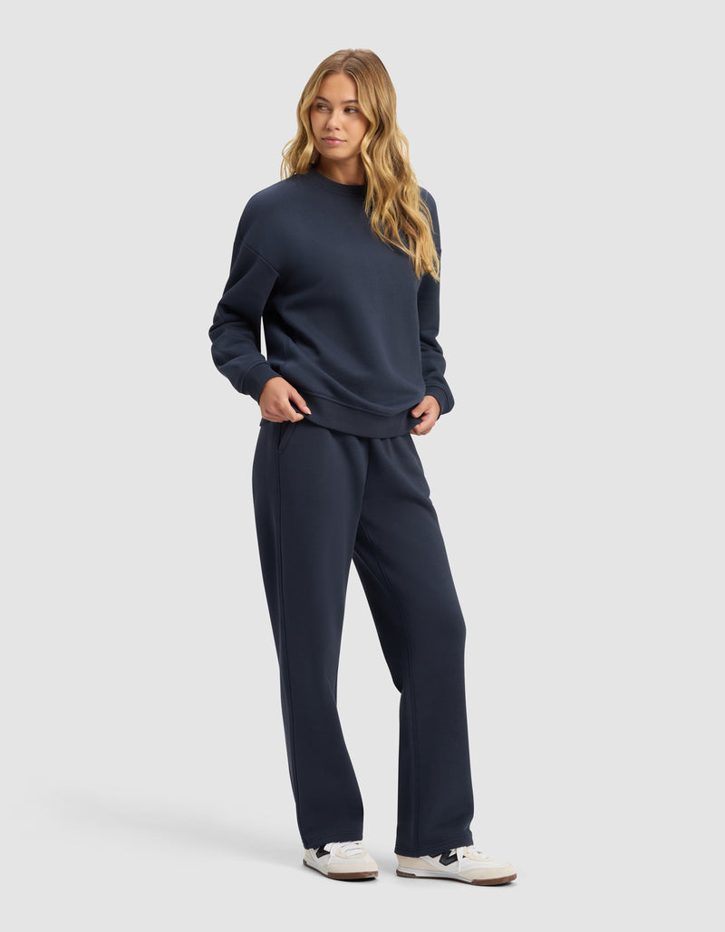 A woman with long, wavy blonde hair models the Cozy Earth Women's Cityscape 2.0 Crewneck & Wide Leg Pant Set in navy blue with white sneakers, standing against a plain light gray background.