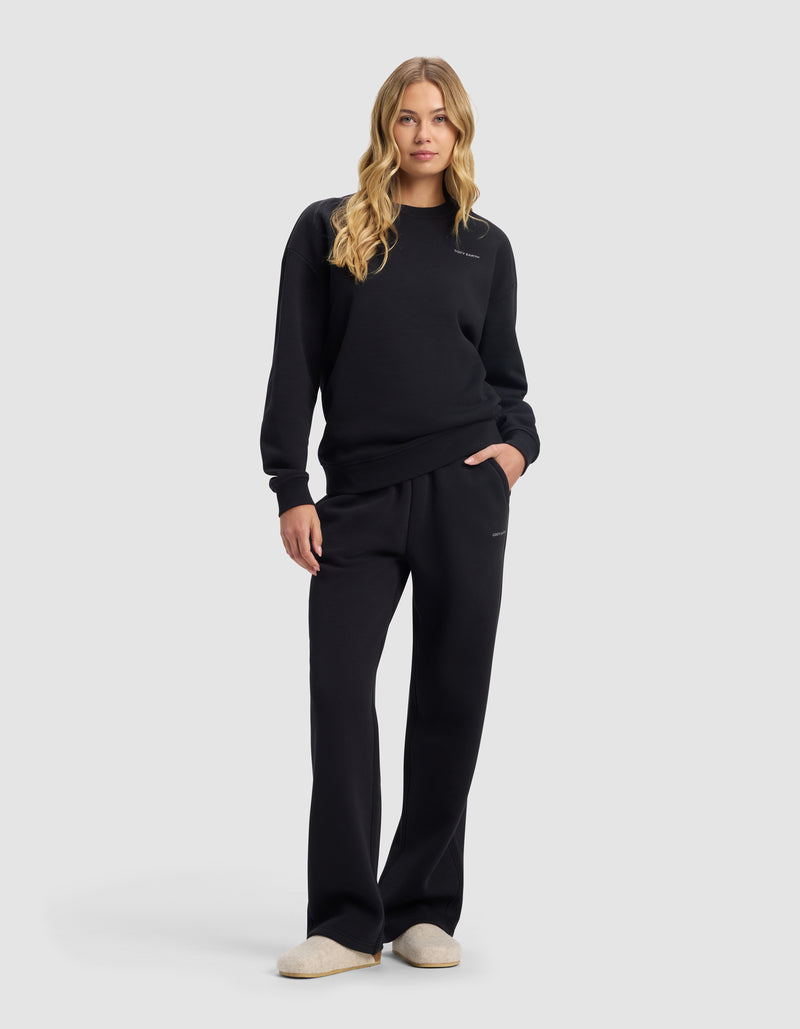 A woman with long blonde hair wears the Cozy Earth Women's Cityscape 2.0 Crewneck & Wide Leg Pant Set in black, standing forward with one hand in her pocket and light-colored slippers, against a plain light background.