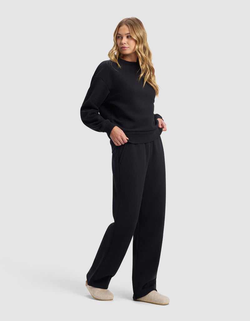 A woman with long wavy blonde hair wears the Cozy Earth Women's Cityscape 2.0 Crewneck & Wide Leg Pant Set in black with light beige slippers, standing against a plain light gray background.