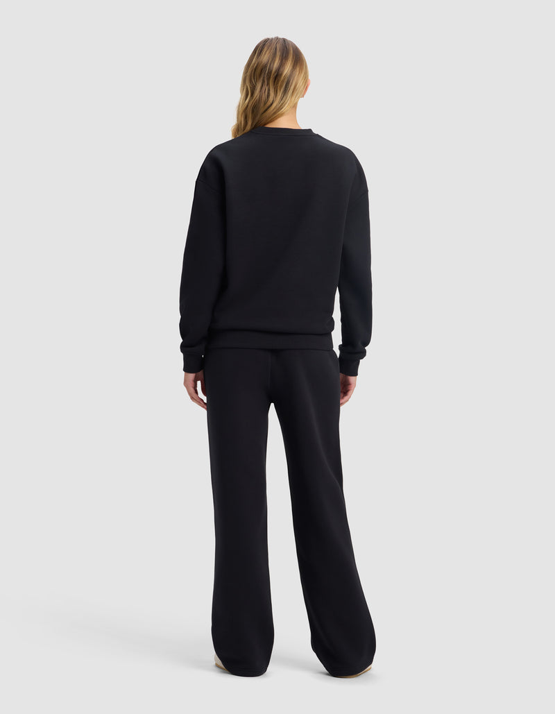 A person with long blonde hair, seen from behind, is wearing the Cozy Earth Women's Cityscape 2.0 Crewneck & Wide Leg Pant Set in black, standing against a plain light gray background.