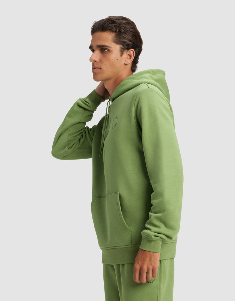 A young man in profile wears the Cozy Earth Men's Cityscape 2.0 Hoodie in light green, paired with matching joggers. His left hand touches the back of his neck against a plain light gray background.