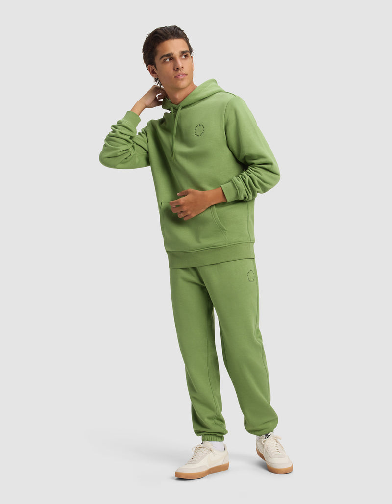 A young man in the Cozy Earth Men's Cityscape 2.0 Hoodie and matching sweatpants with white sneakers stands against a plain background, looking to the side with one hand touching his neck.