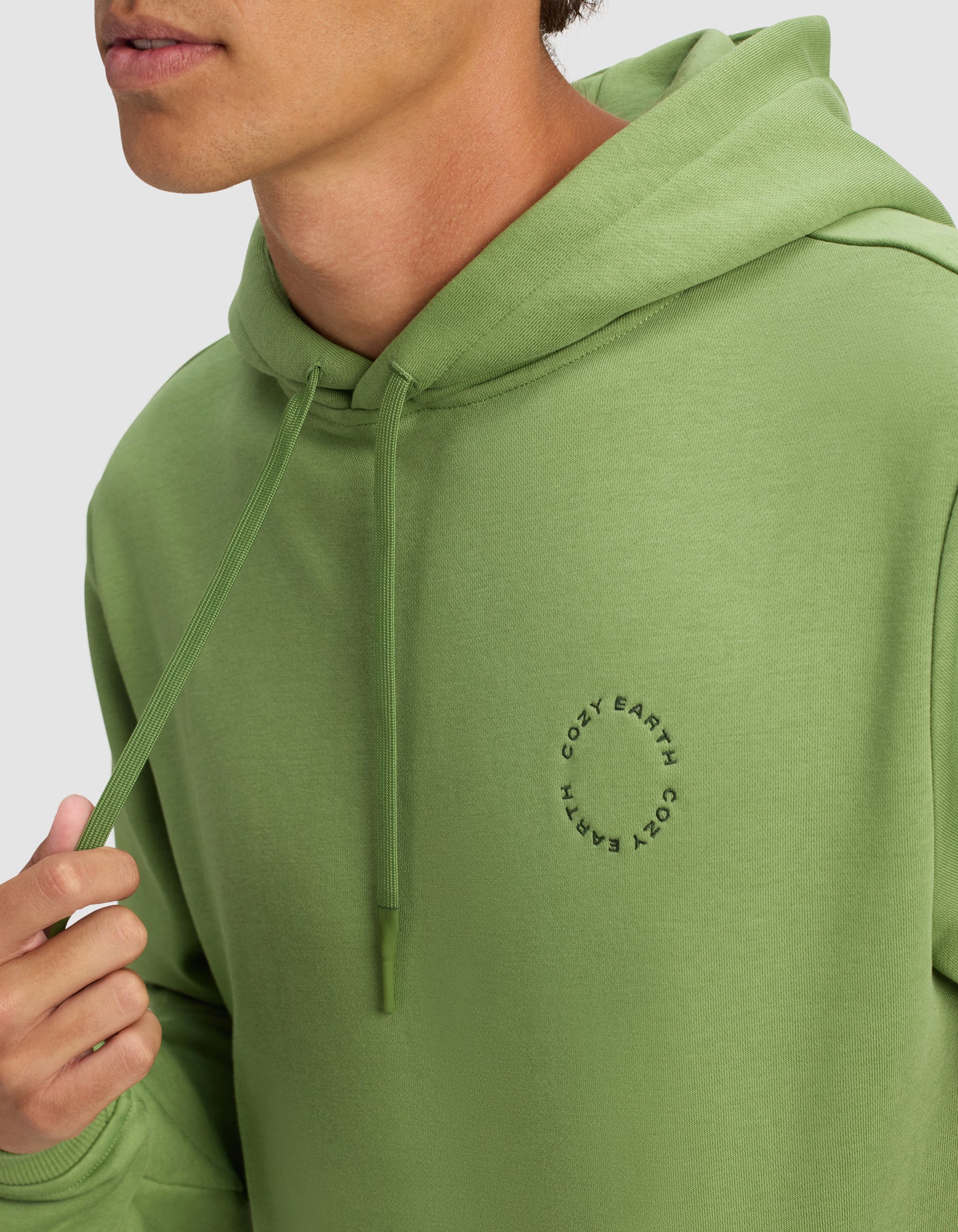 A man wears the Men's Cityscape 2.0 Hoodie by Cozy Earth in light green, featuring a small circular logo on the chest. He holds a hoodie drawstring, with only his lower face visible.