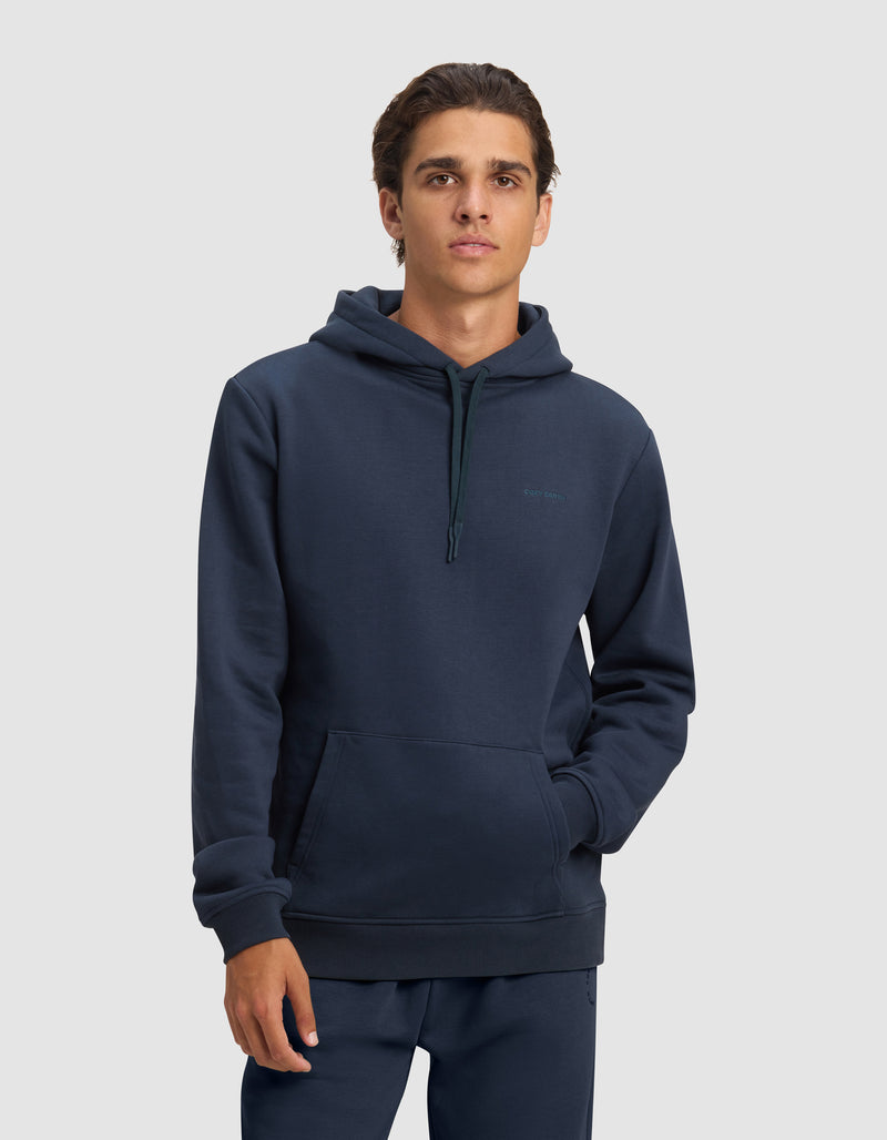A young man with short brown hair models the Cozy Earth Men's Cityscape 2.0 Hoodie in dark blue, featuring a front pocket and hood, paired with matching pants against a light gray background.