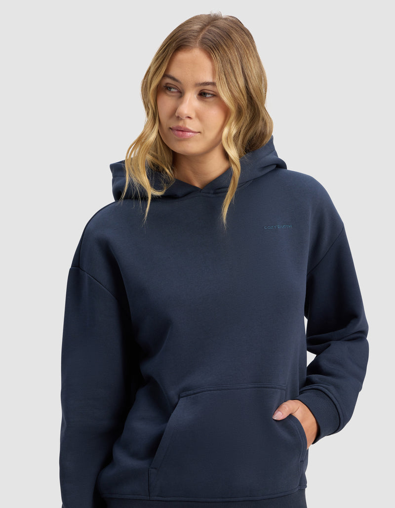 A woman with long, wavy blonde hair wears the Cozy Earth Women's Cityscape 2.0 Hoodie in dark navy, with one hand in the pocket, gazing slightly to the side against a plain light gray background.