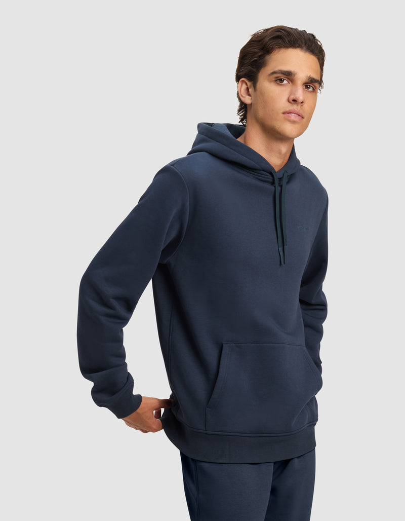 A young man models the Cozy Earth Men's Cityscape 2.0 Hoodie in dark blue with matching pants, standing with one hand behind his back and gazing to the side against a plain light gray background.