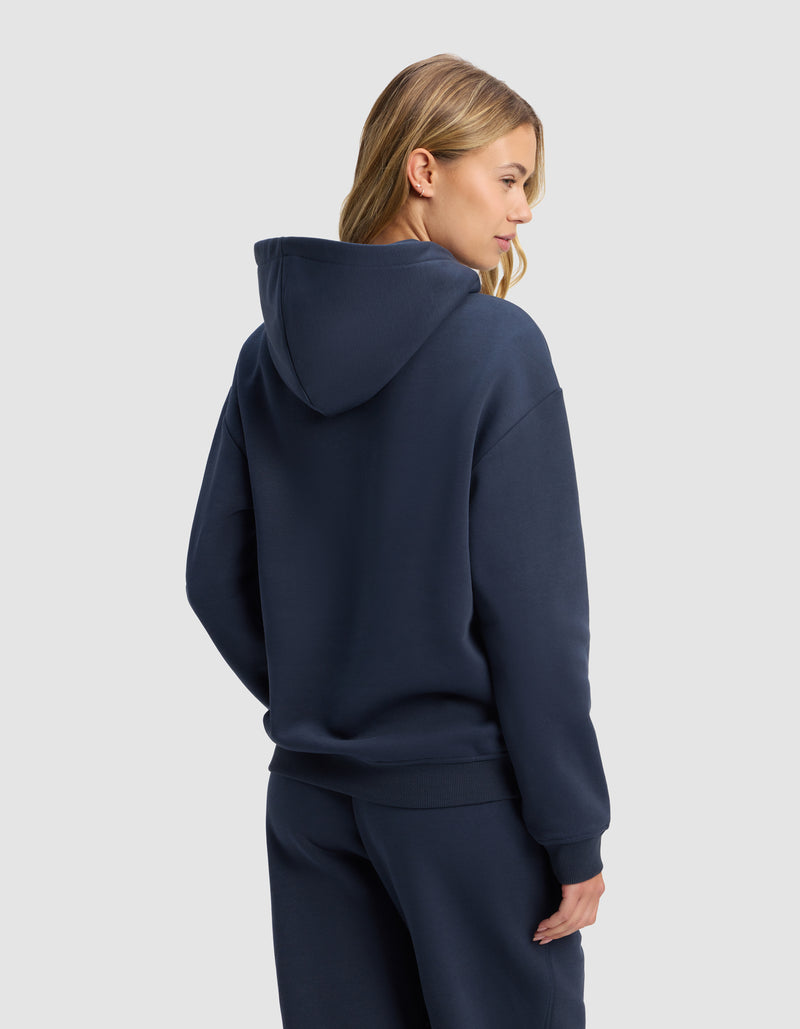 A woman stands with her back and side to the camera against a plain light gray background, wearing the Cozy Earth Women's Cityscape 2.0 Hoodie in navy blue with matching sweatpants. Her long blonde hair is visible.
