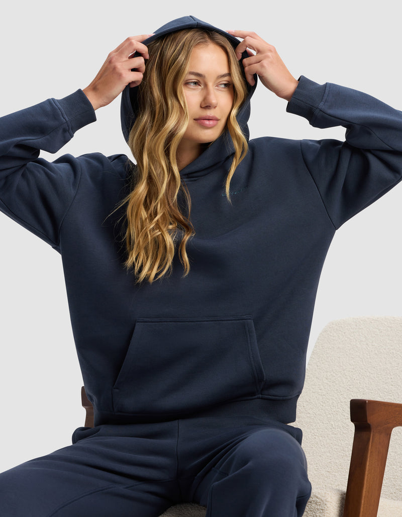 A woman with long blonde hair sits on a light chair, wearing the Cozy Earth Women's Cityscape 2.0 Hoodie in dark blue and matching sweatpants, adjusting the hood with both hands while looking slightly to the side.