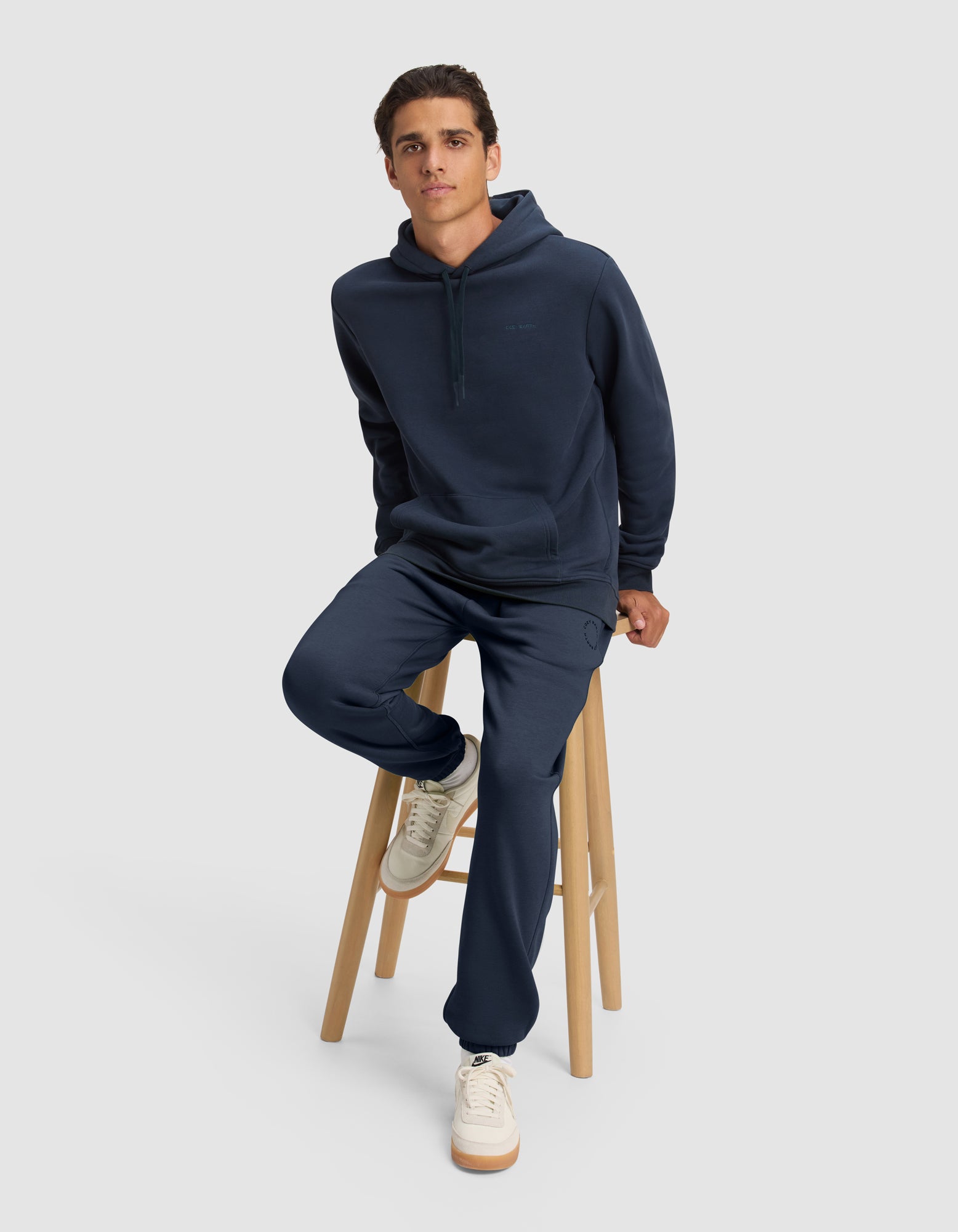 A man wears the Cozy Earth Men's Cityscape 2.0 Hoodie and matching sweatpants, sitting on a wooden stool against a plain light gray background, with one foot on the stool’s lower rung and white sneakers visible.