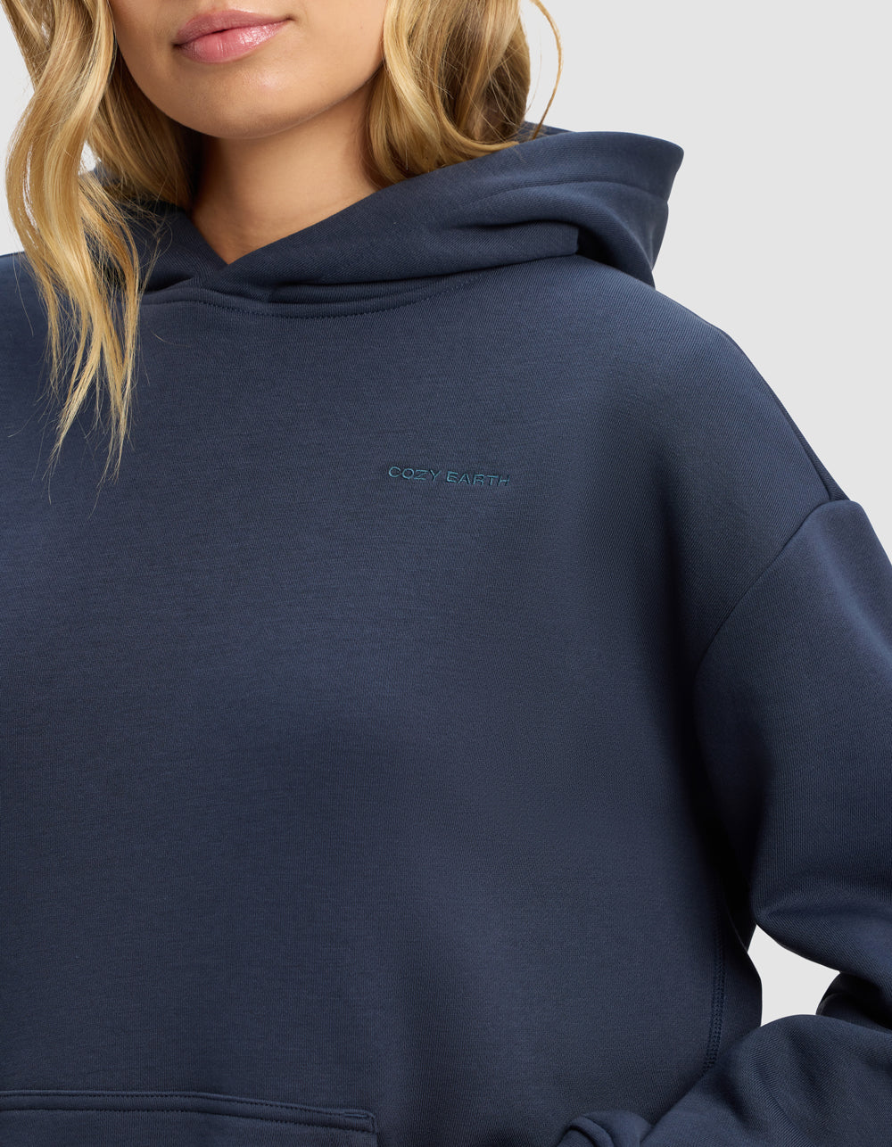 A woman wears the Cozy Earth Women's Cityscape 2.0 Hoodie in navy blue, featuring a small embroidered logo on the chest. Only her lower face and upper body appear against a plain background.