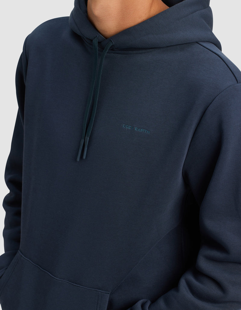 A person wears the Men's Cityscape 2.0 Hoodie by Cozy Earth—a dark blue hoodie with a front pocket, drawstrings, and "COS EARTH" embroidered on the chest. The person’s face is not shown.