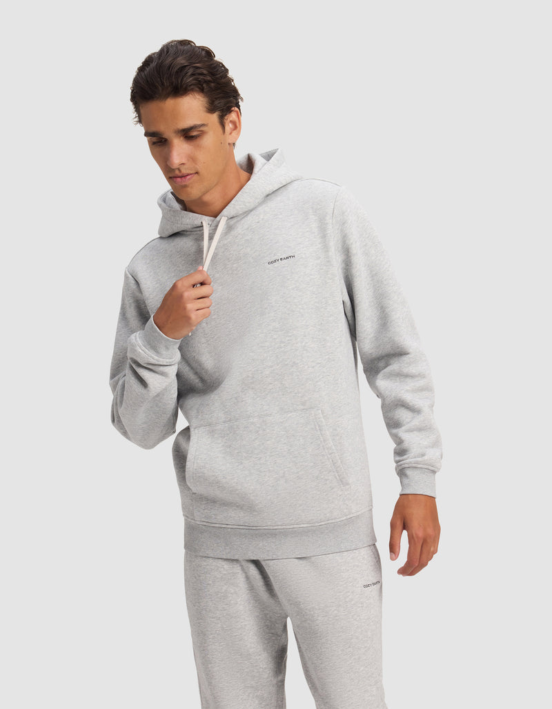 A man wears the Cozy Earth Men's Cityscape 2.0 Hoodie in light gray, paired with matching sweatpants, standing against a plain background while holding a drawstring and looking down with a relaxed expression.