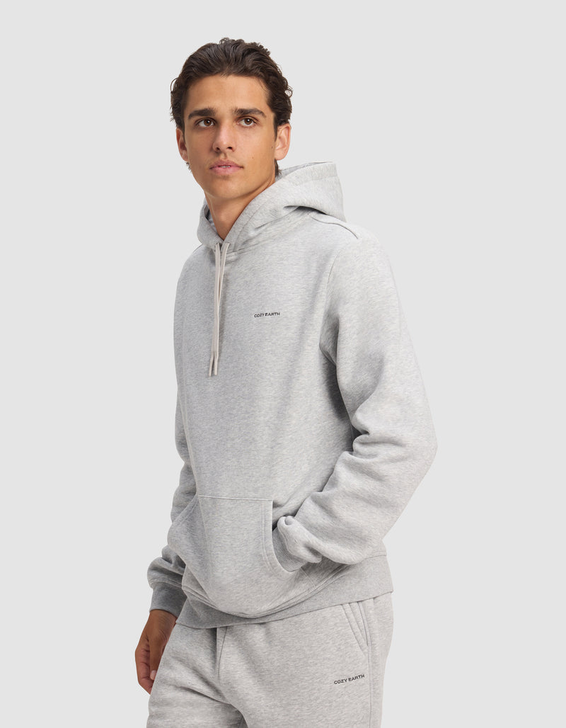 A young man models the Cozy Earth Men's Cityscape 2.0 Hoodie in light gray, with matching sweatpants, standing against a plain background and gazing slightly to the side with one hand in his pocket.