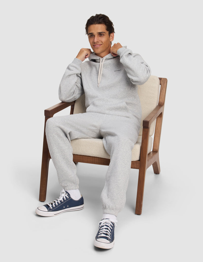 A young man relaxes on a cushioned chair, smiling slightly as he holds the drawstrings of his Cozy Earth Men's Cityscape 2.0 Hoodie in light gray, paired with sweatpants and dark blue sneakers.